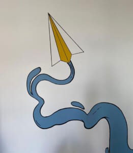 Dettaglio dell'aeroplanino di carta con scia blu dipinto sul muro